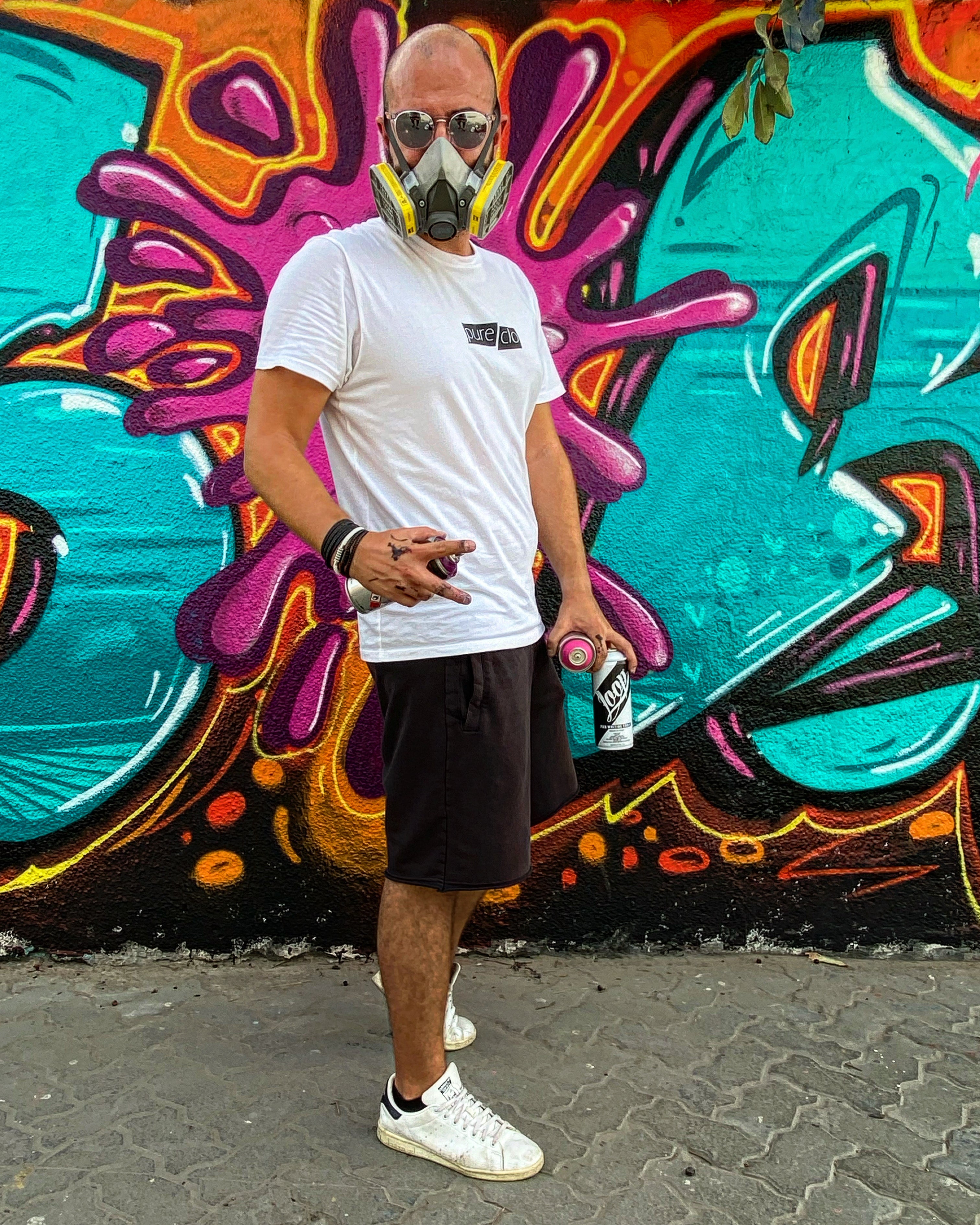Spam wearing a white t-shirt and black shorts standing in front of a colorful graffiti wall.