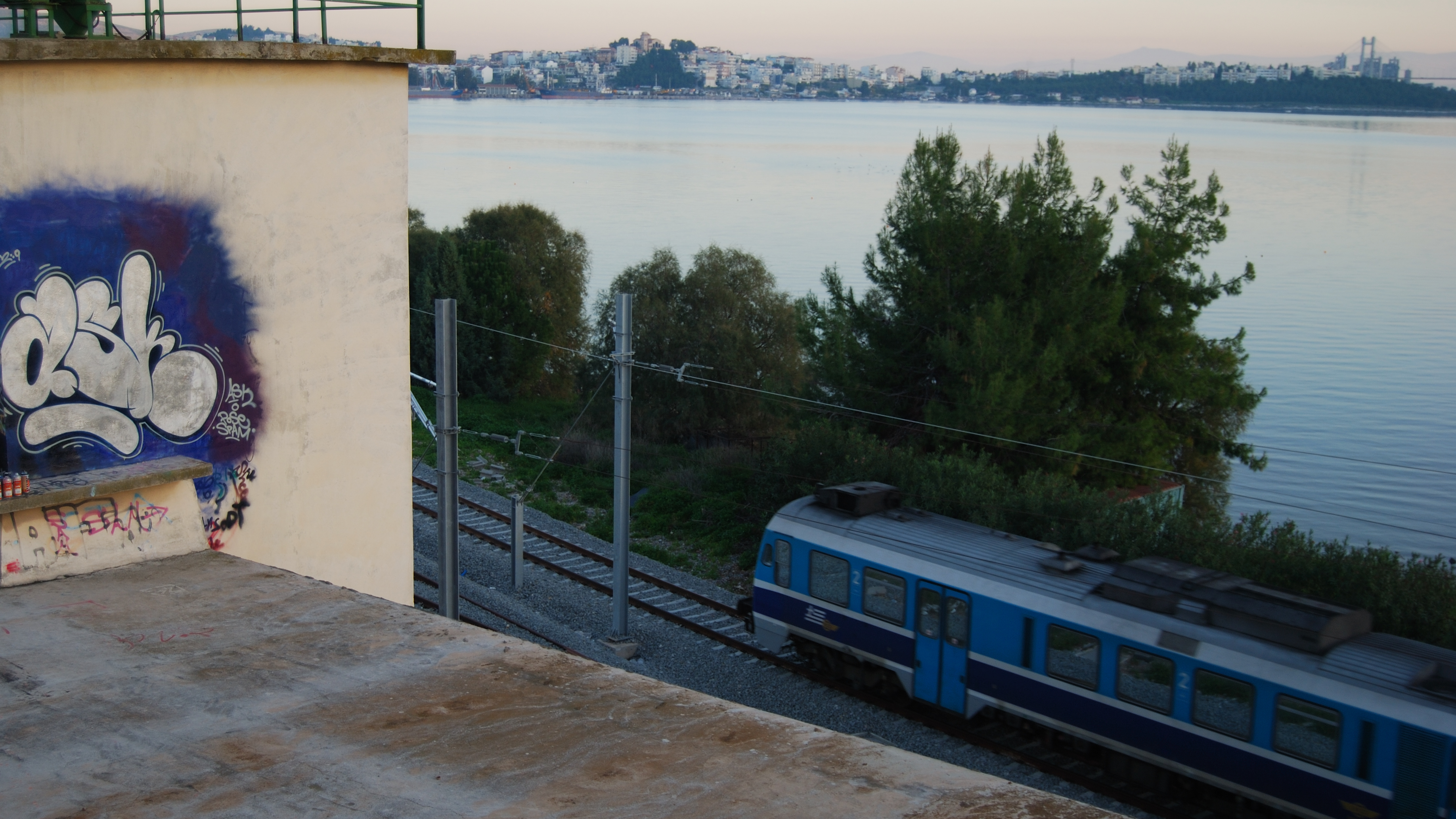 Train passing by a waterfront with a graffiti on a wall and trees in the background